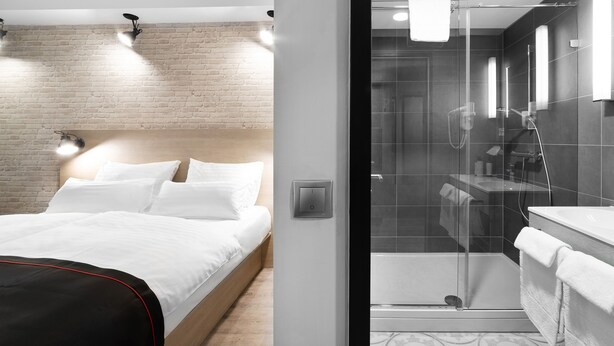 Una habitación de hotel moderna con pared de ladrillo decorativa, cama con ropa blanca, una silla negra y cabina de ducha de vidrio con una toalla blanca colgada en la puerta.