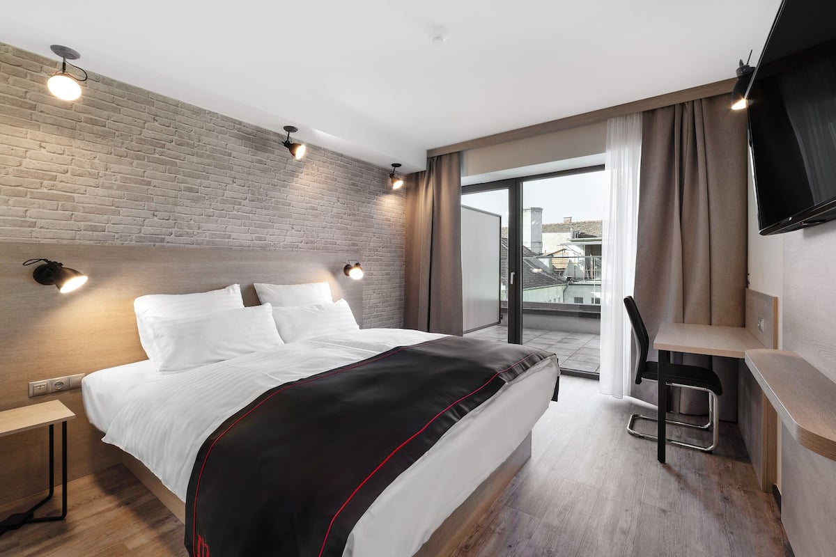 A modern hotel room with a brick accent wall, a large bed with white bedding, a desk with a chair, and a TV mounted on the wall.