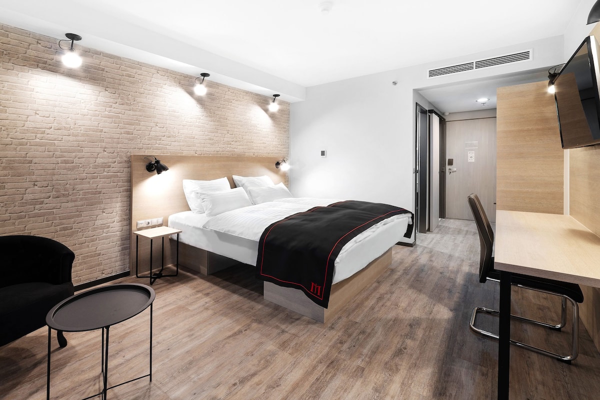 A modern hotel room with a brick wall, a bed with white bedding, a small table with a chair, and a desk with a chair.