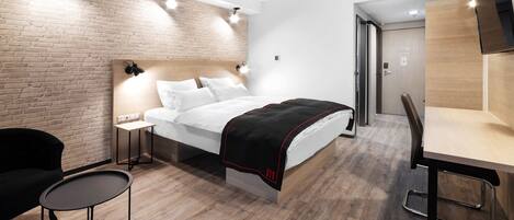 A modern hotel room with a brick wall, a bed with white bedding, a small table with a chair, and a desk with a chair.