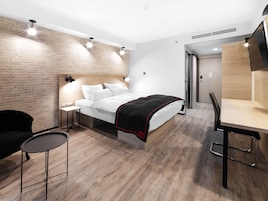 A modern hotel room with a brick wall, a bed with white bedding, a small table with a chair, and a desk with a chair.