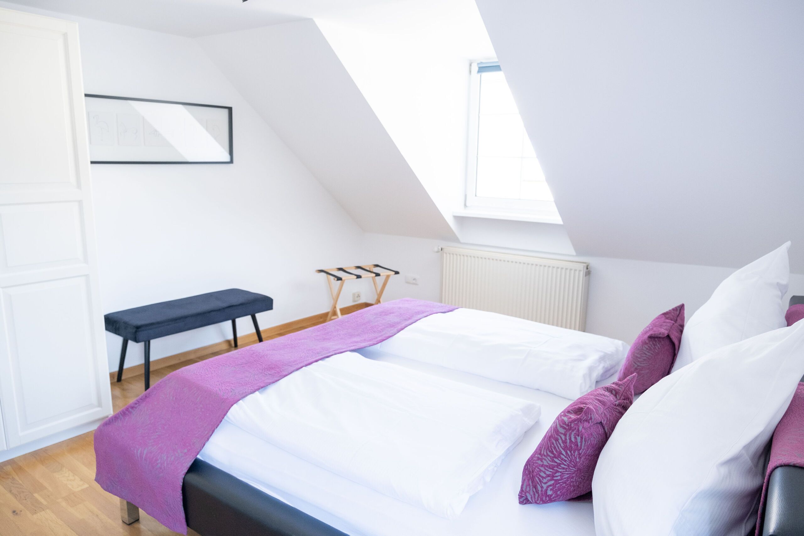 Un dormitorio moderno con techo inclinado, cama con ropa de cama blanca y detalles morados, un banco y un cuadro en la pared.