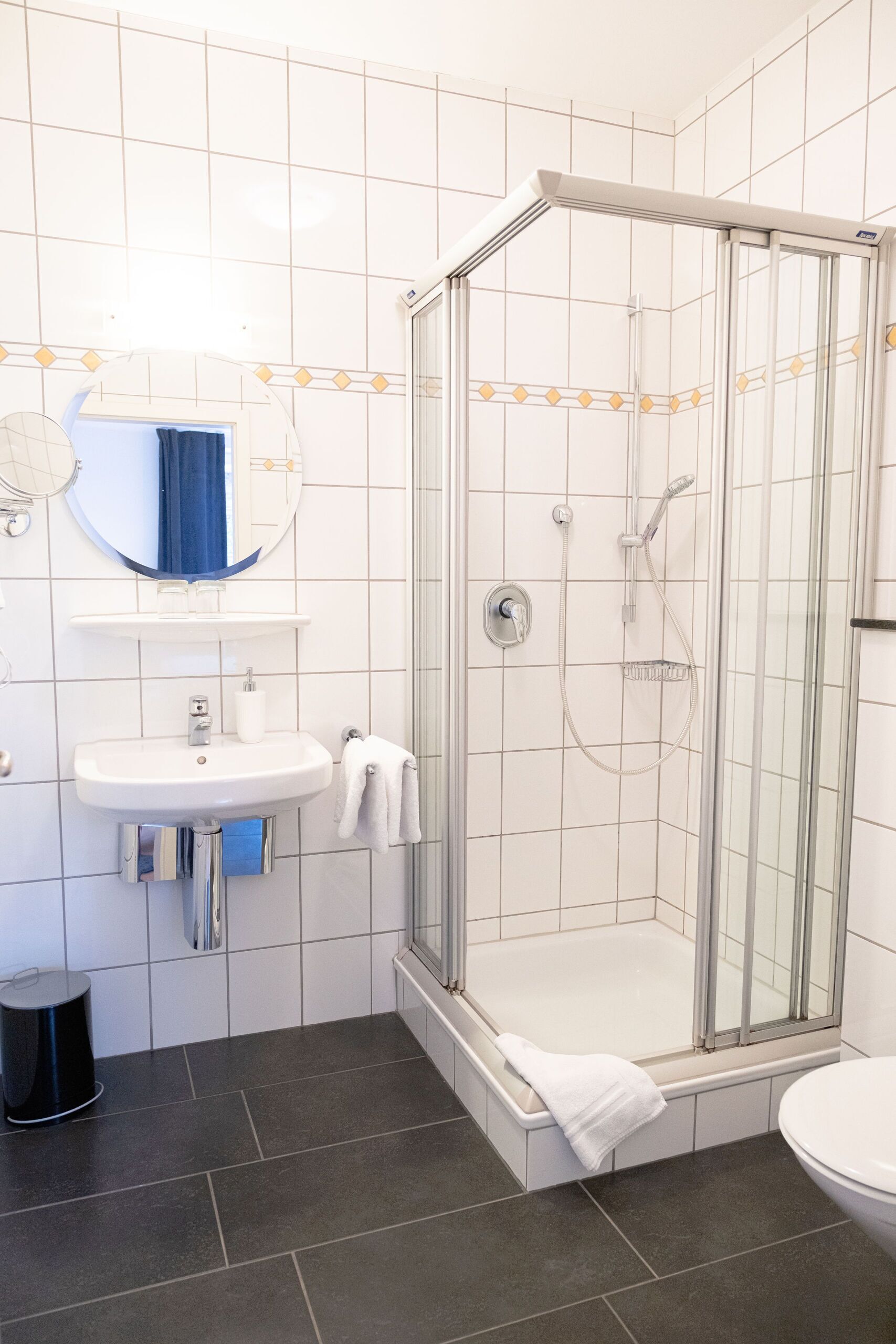 A modern bathroom with a glass-enclosed shower, a white sink with a mirror above it, and a black trash bin.