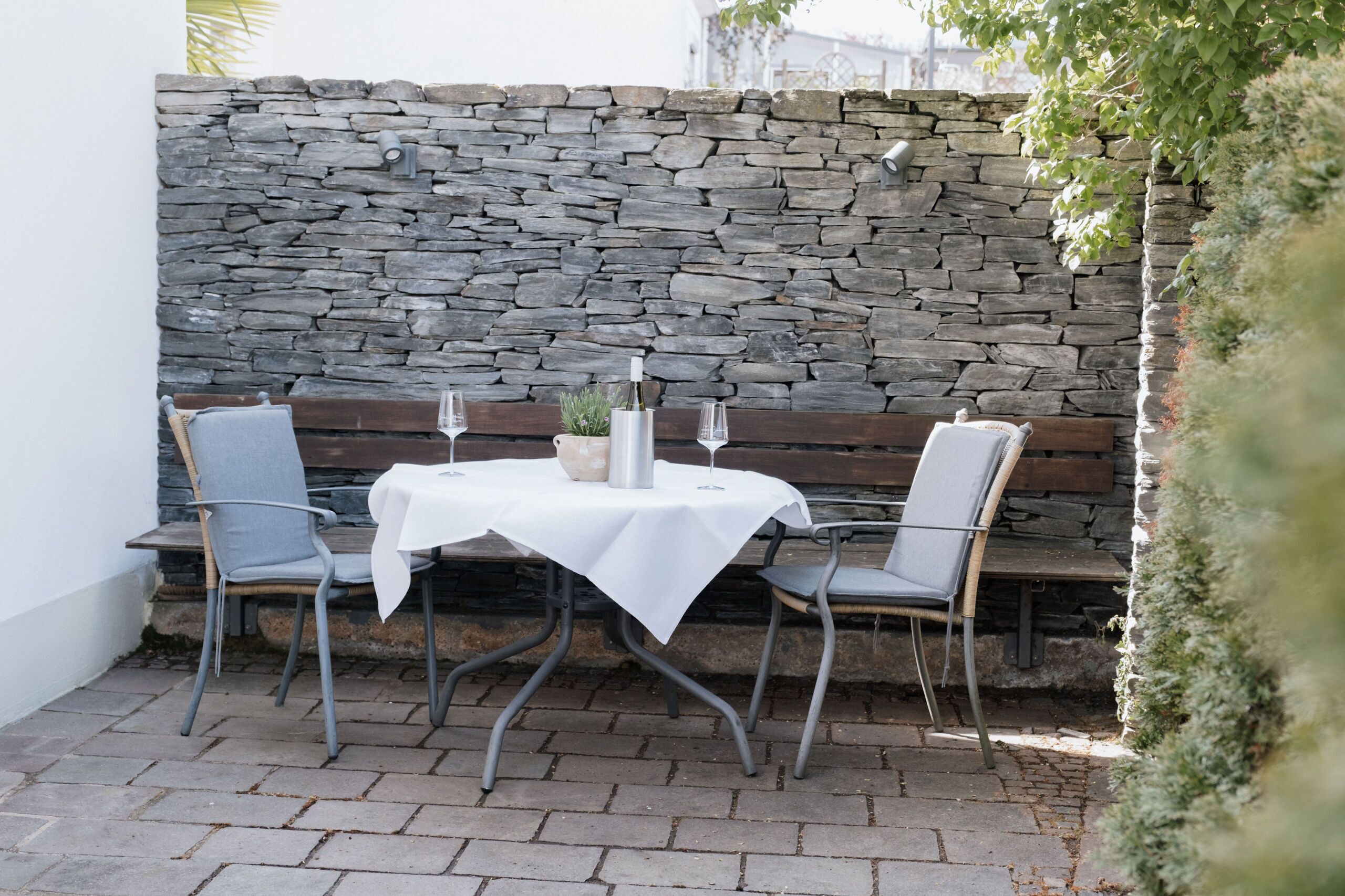 Een terras buiten met een muur van natuursteen, een ronde tafel met een witte tafelkleed en twee stoelen.