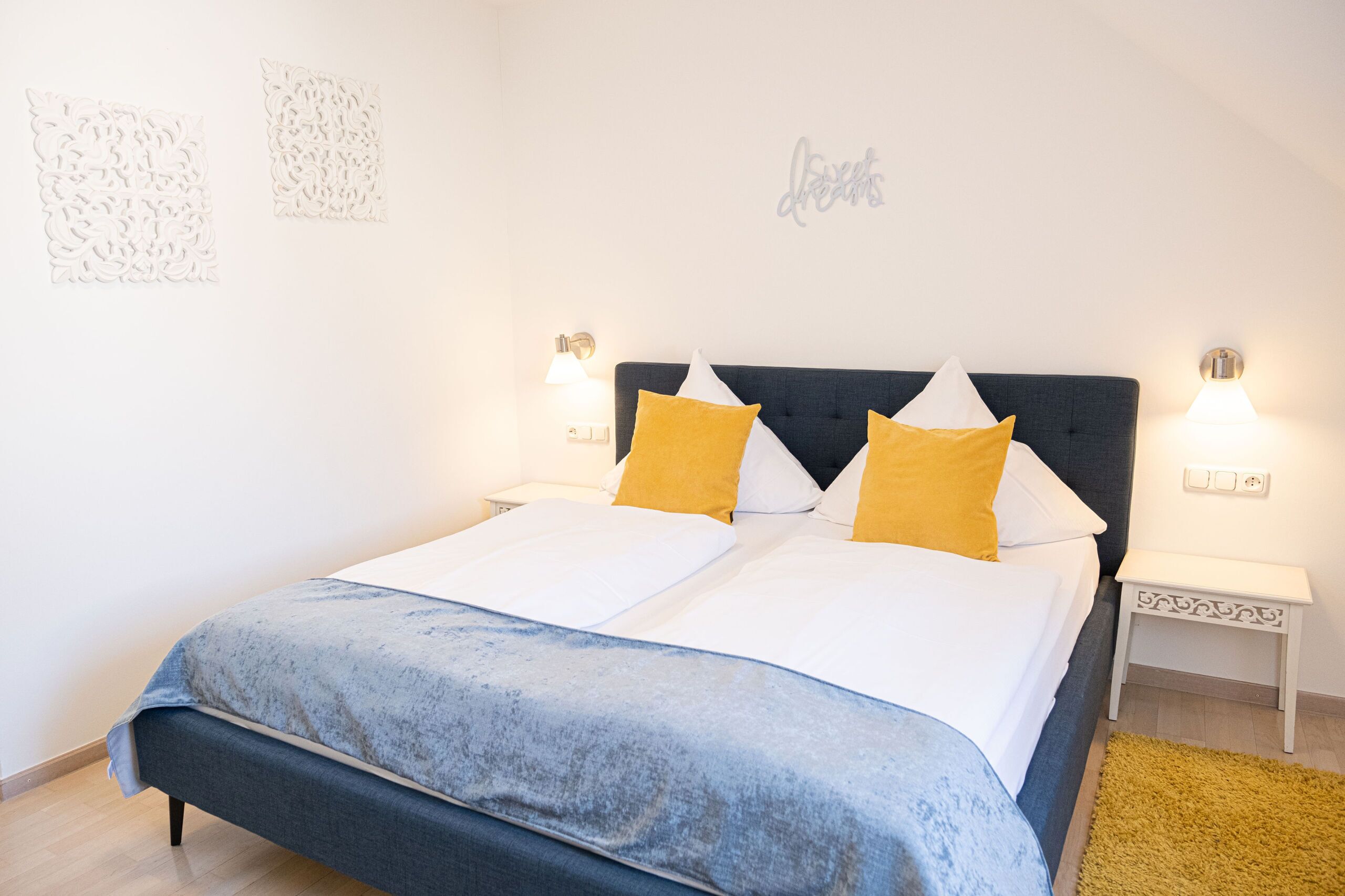Una cama bien tendida con un cabecero azul, dos mesitas de noche con lámparas y un adorno en la pared.