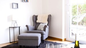 A modern living room with a grey armchair, a small side table with a lamp, a dark rug, and a glass table with wine glasses and a bottle. - Cabinett 1876 (Trittenheim)