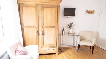 A room with a wooden wardrobe, a glass table with a vase, a white armchair, and a television on the wall.