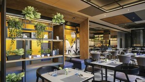 A modern restaurant interior with multiple dining tables and chairs, wooden shelves with potted plants, and a kitchen area in the background. - ibis Surabaya Tidar (Surabaya)