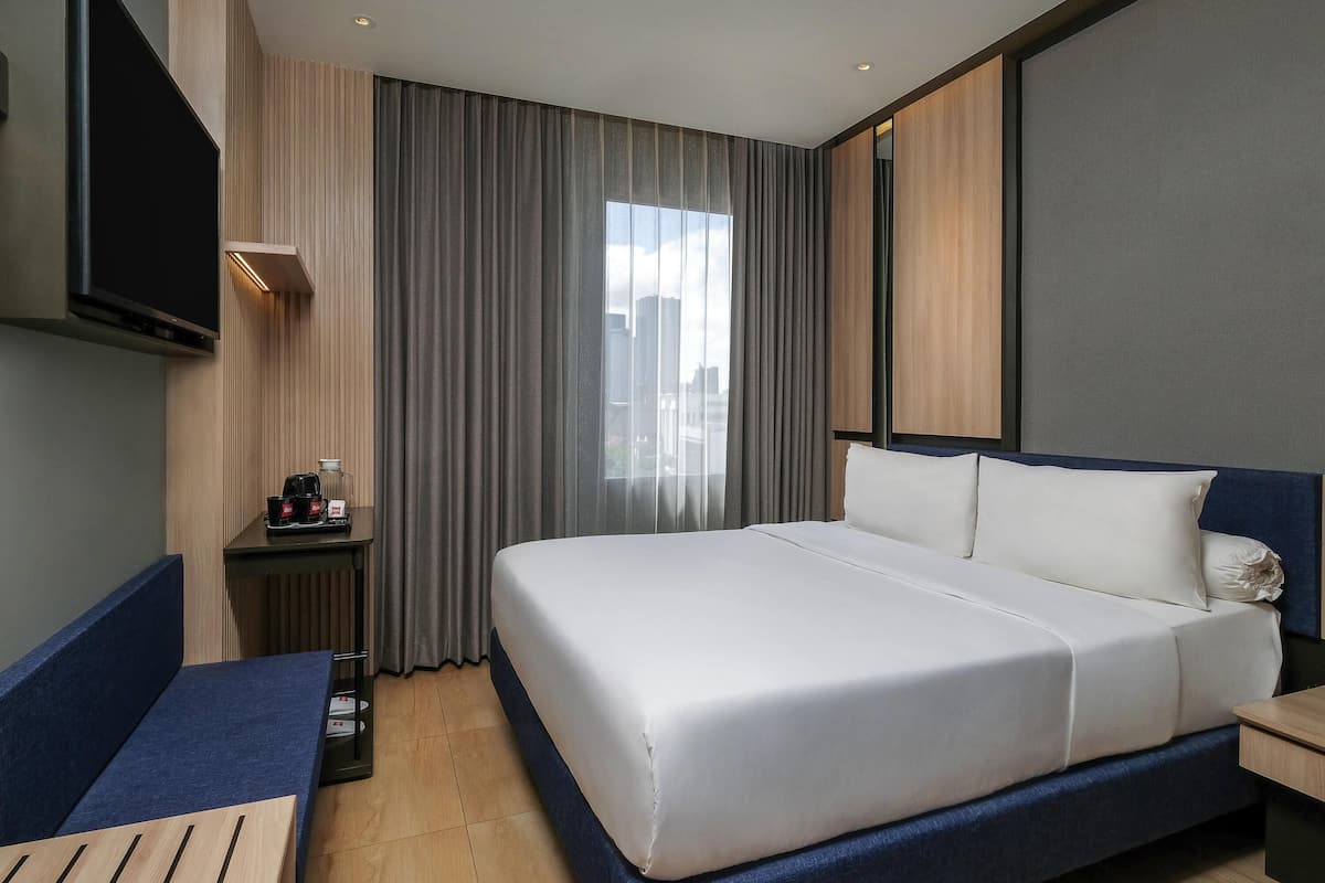 A modern hotel room with a large bed, a flat-screen TV mounted on the wall, a small table with a coffee maker, and a view of the city through the window.