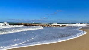 Una platja amb onades que trenquen a la riba, un moll de pedra i un cel blau clar.