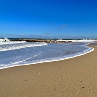 Una playa con olas rompiendo en la orilla, un rompeolas rocoso y un cielo azul despejado.