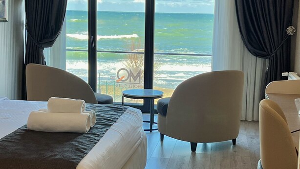 Habitació d'hotel amb un llit gran, dues butaques, una taula petita i vista a l'oceà a través de grans finestres.