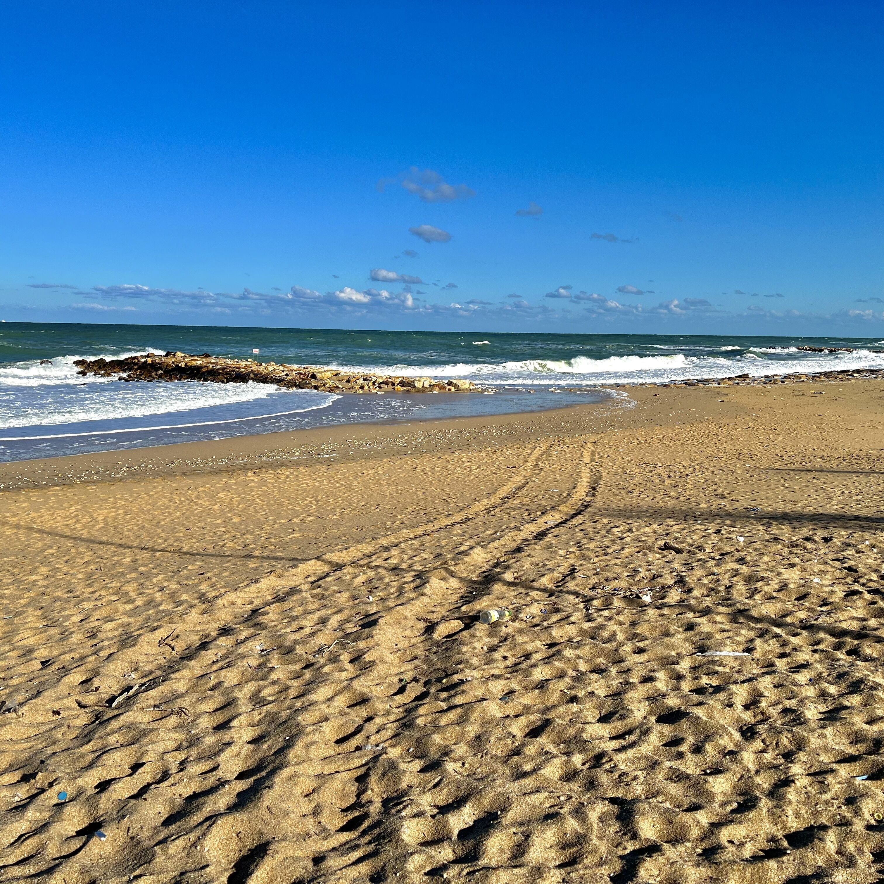 Una playa arenosa con huellas que conducen al agua, un rompeolas rocoso y un cielo azul despejado.