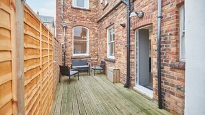 A narrow wooden deck with a brick building on one side, a small table with chairs, and a door on the other.