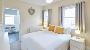 A bedroom with a large bed, two bedside tables, a nightstand, a radiator, a window with curtains, and a wall-mounted wreath.