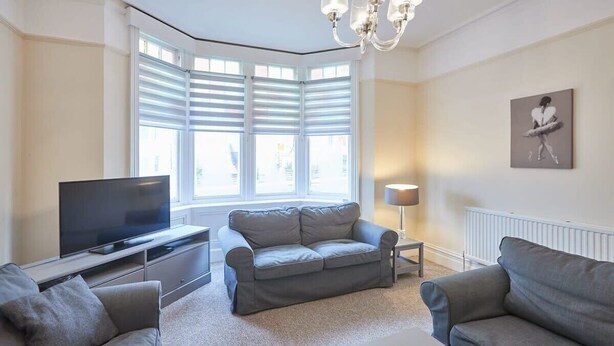 A living room with a flat-screen TV, grey sofas, a coffee table, and a large window with blinds.