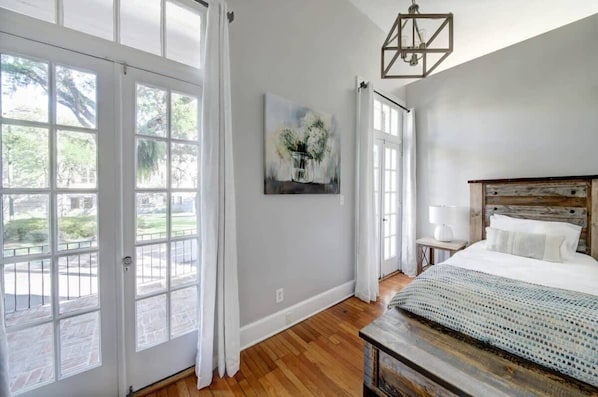 A bedroom with a large window, a bed, a nightstand, and a painting on the wall.