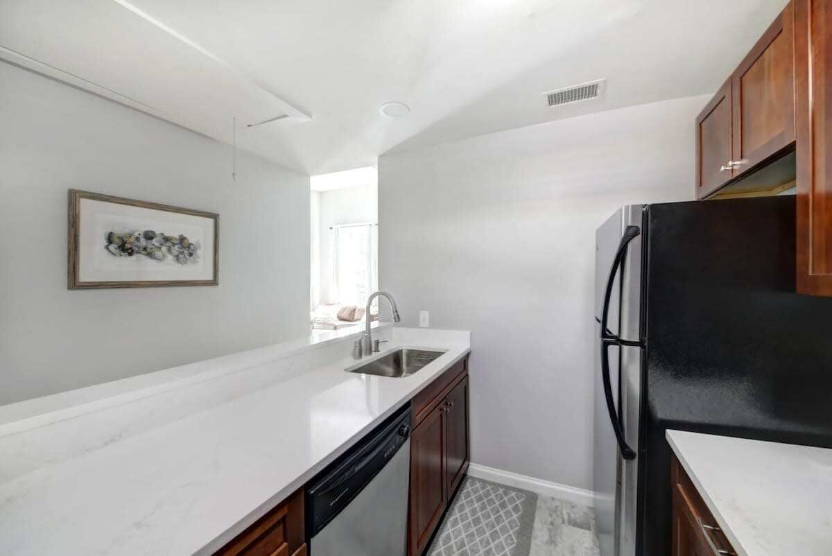 Una cocina con mostradores blancos, lavavajillas, refrigerador, armarios de madera y una ventana.
