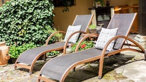 Drie houten lounge stoelen met grijze kussens en gestreepte kussens, geplaatst op een stenen terras met een weelderige groene haag op de achtergrond.