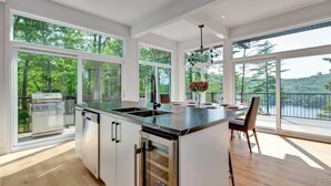 Une cuisine moderne dotée d’un grand îlot central, d’un lave-vaisselle et d’un barbecue, avec de grandes fenêtres offrant une vue sur l’extérieur.