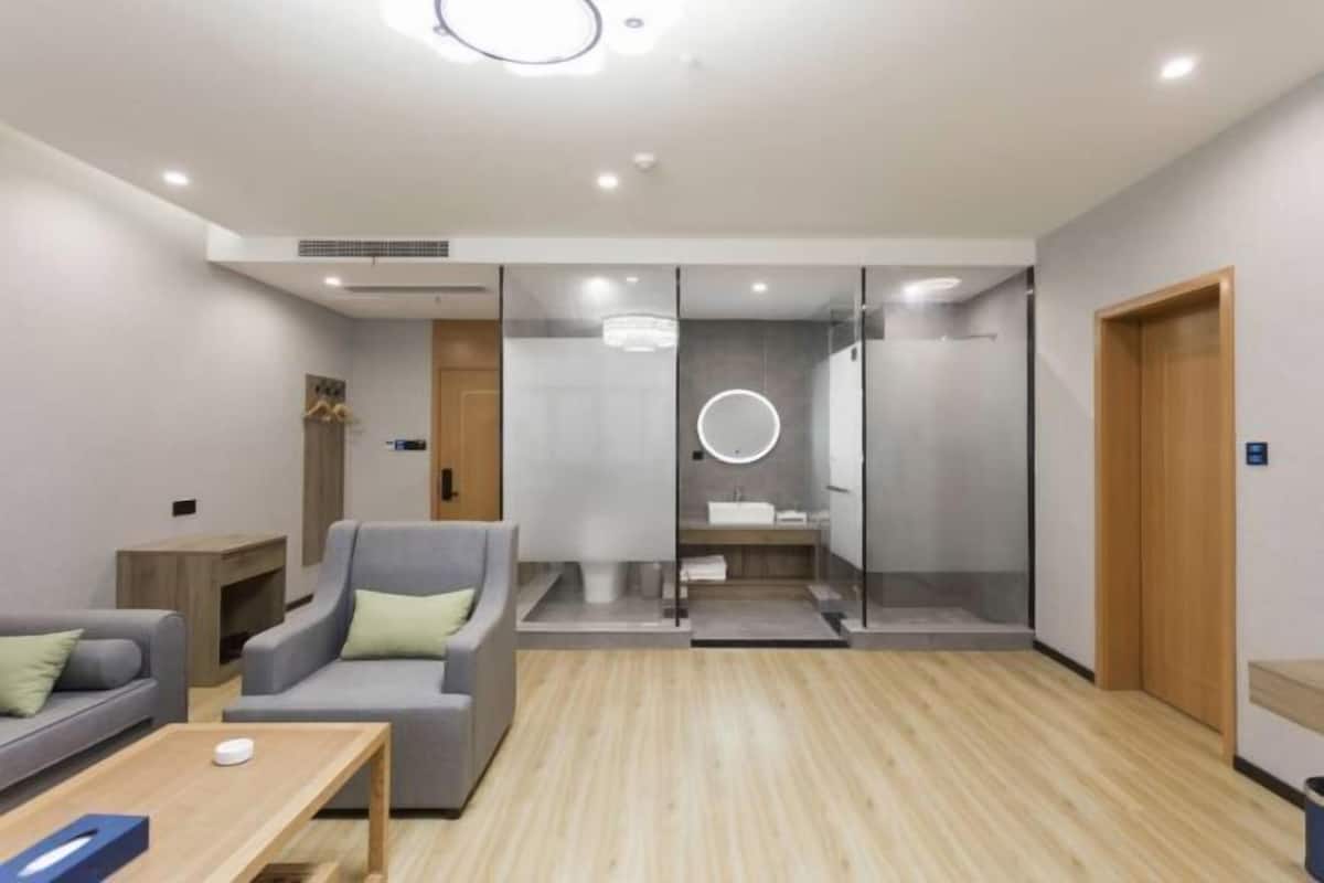 Una habitación de hotel moderna con un sofá gris, una mesa de centro de madera y un baño con paredes de vidrio.
