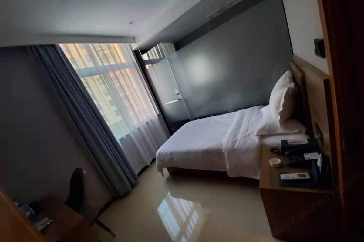 Habitación de hotel con cama, mesita de noche con teléfono y control remoto, ventana con cortinas y radiador.
