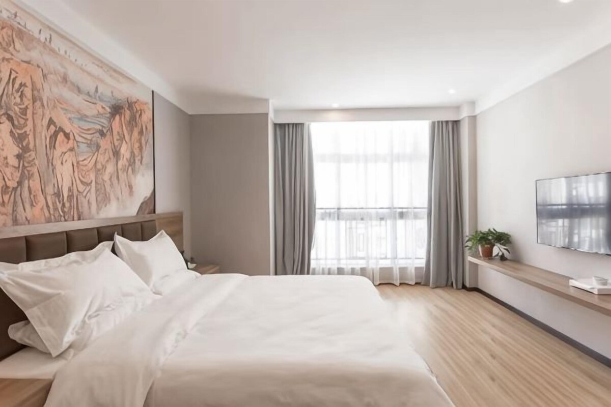A modern hotel room with a large bed, a painting on the wall, a window with curtains, and a small plant on a shelf.