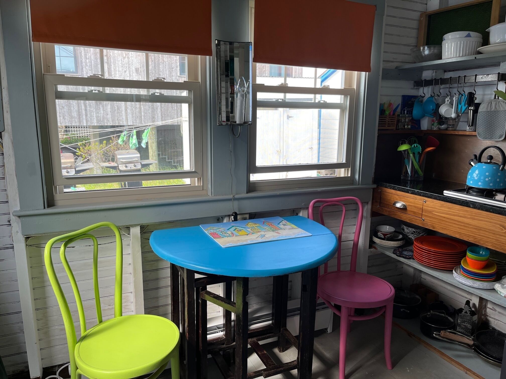 Une cuisine avec une table bleue, deux chaises, une chaise rose et une fenêtre avec des stores.