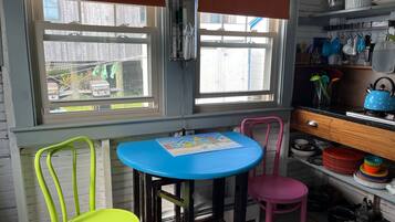 Une cuisine avec une table bleue, deux chaises, une chaise rose et une fenêtre avec des stores.
