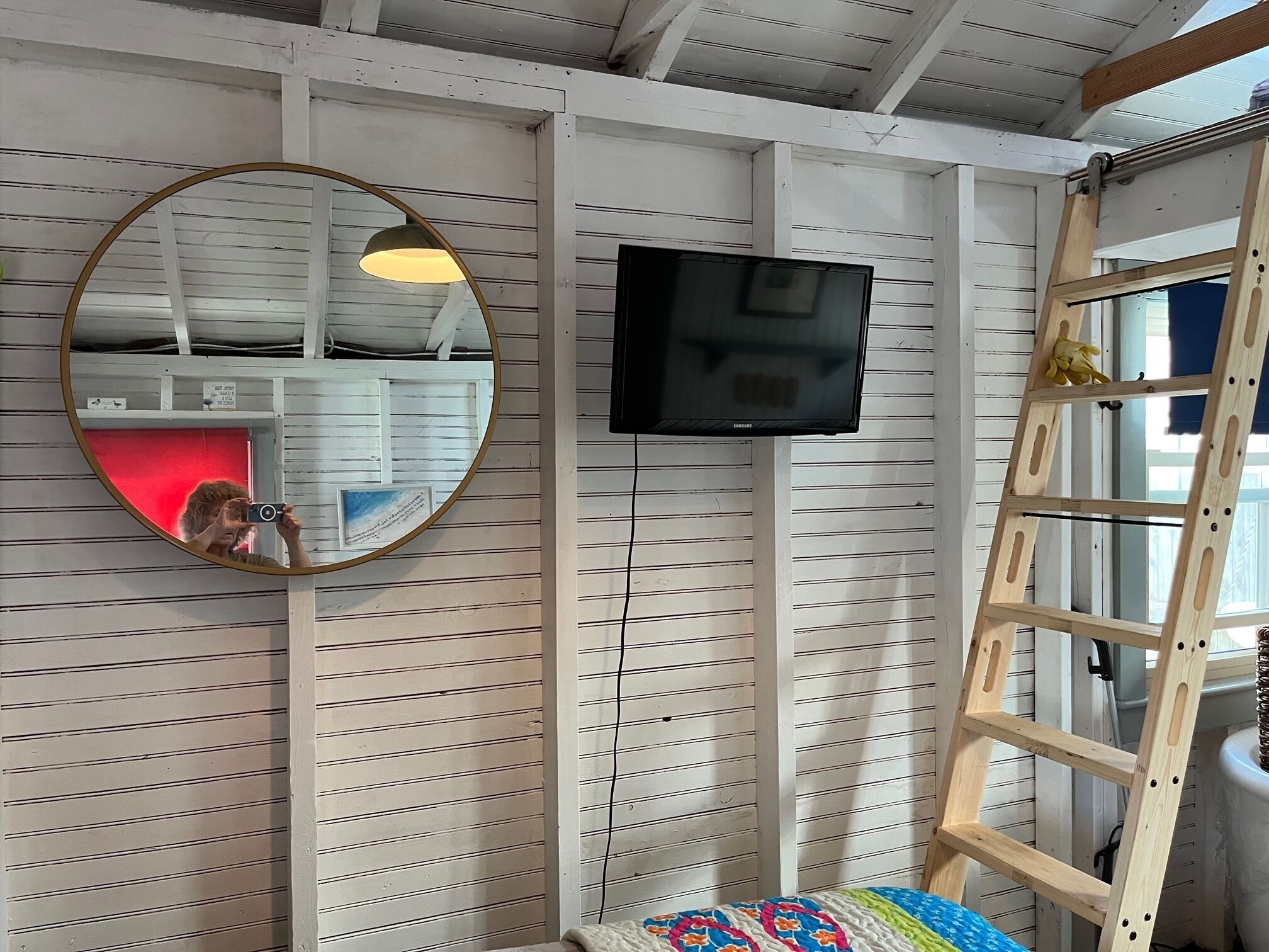 A room with a white paneled wall, a round mirror, a flat-screen TV, and a wooden ladder.