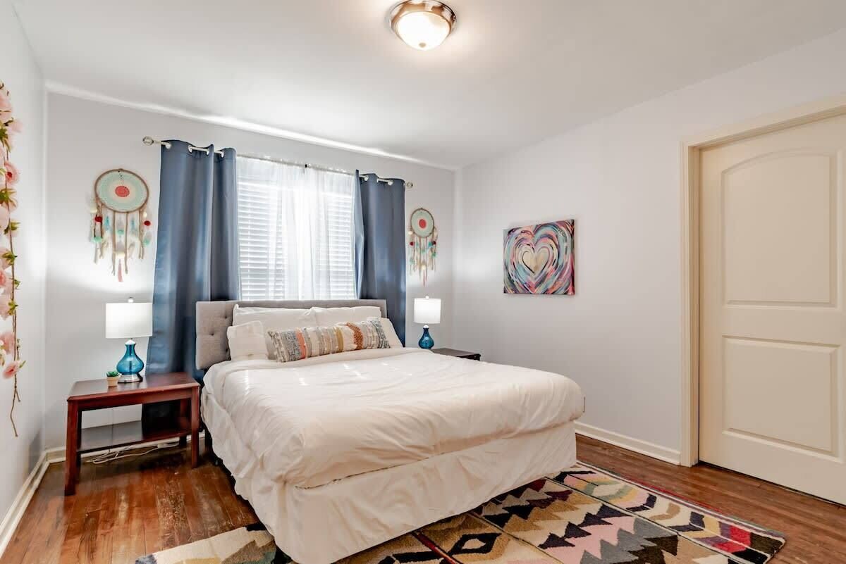 Een slaapkamer met een bed, twee nachtkastjes, gordijnen voor het raam, een dromenvanger en een decoratief kunstwerk aan de muur.