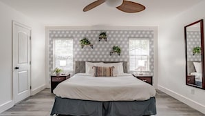 Un dormitorio con una cama grande, dos mesitas de noche, un ventilador de techo y tres estantes en la pared con plantas.