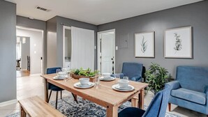 Comedor con una mesa de madera dispuesta para cuatro personas, rodeada de sillas azules. Hay grabados botánicos enmarcados en la pared, una planta en maceta y un sofá azul al fondo.