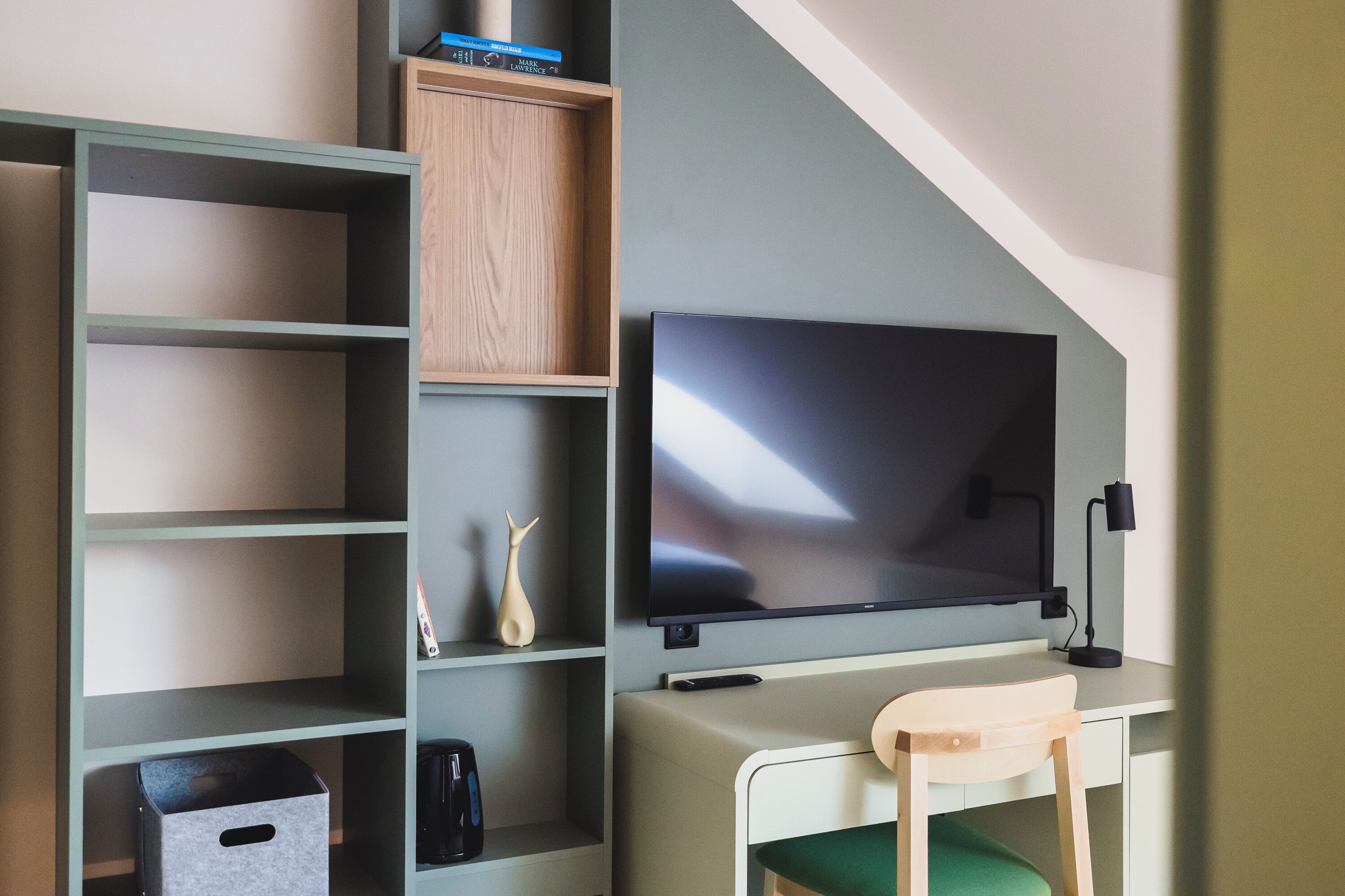 A room with a flat-screen TV mounted on the wall, a desk with a chair, and a shelving unit with various items.