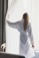 Una persona vestida con una bata blanca, de pie junto a una ventana con cortinas translúcidas, una silla y una mesa de madera al frente.