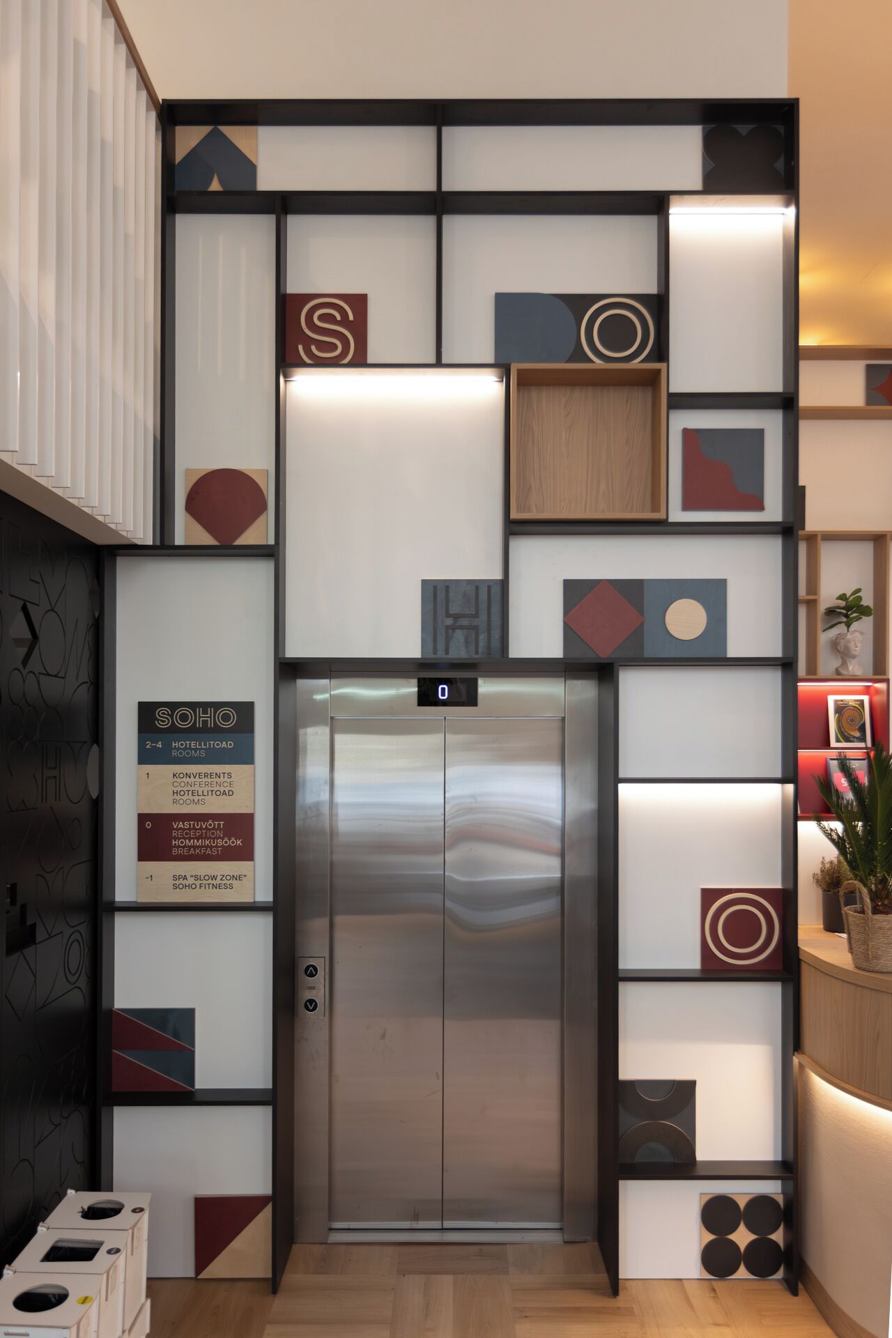 An elevator with a digital display, surrounded by a modern wall with geometric patterns and a sign indicating hotel rooms and amenities.