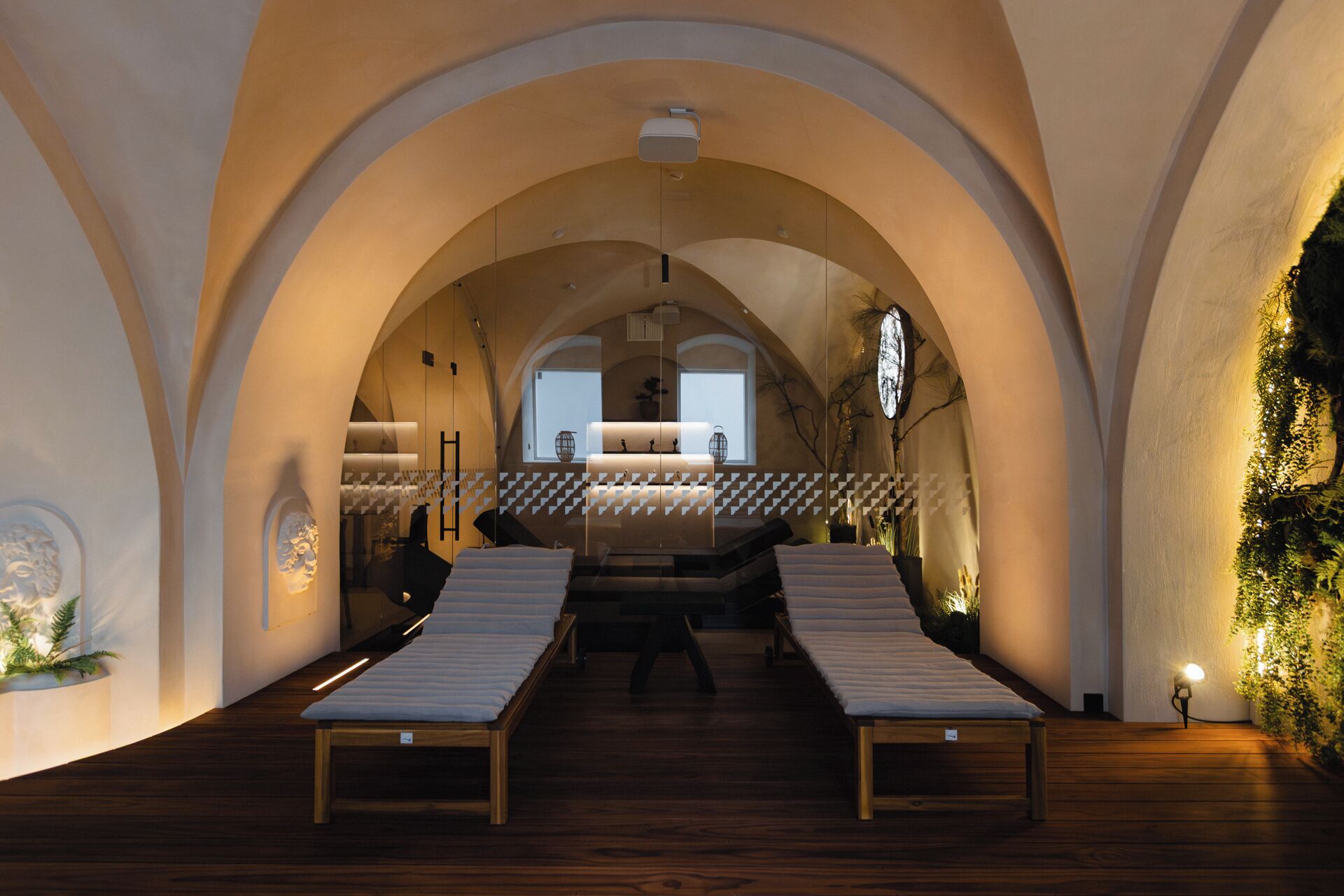 A room with a high arched ceiling, a central bed with a white mattress, and a small table with a lamp.