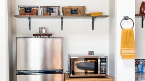 A kitchen with a stainless steel refrigerator, a microwave, and wooden shelves above holding jars and a basket.