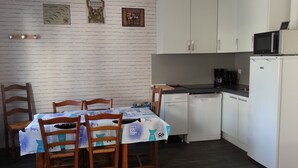Une cuisine compacte avec des meubles blancs, un four à micro-ondes et un lave-vaisselle. On y trouve une table à manger avec des chaises et une nappe ornée de motifs de poissons.