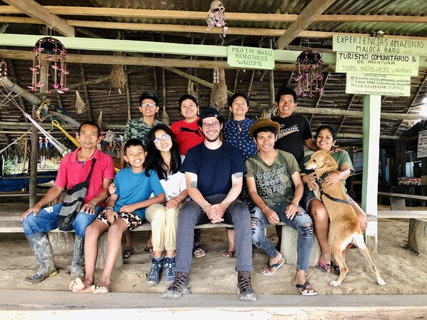 A group of people seated on steps under a structure with hanging decorations and signs indicating a communal tourism experience. - Amazonas Maloca Barü, Hostel Tikuna Macedonia  (Leticia)