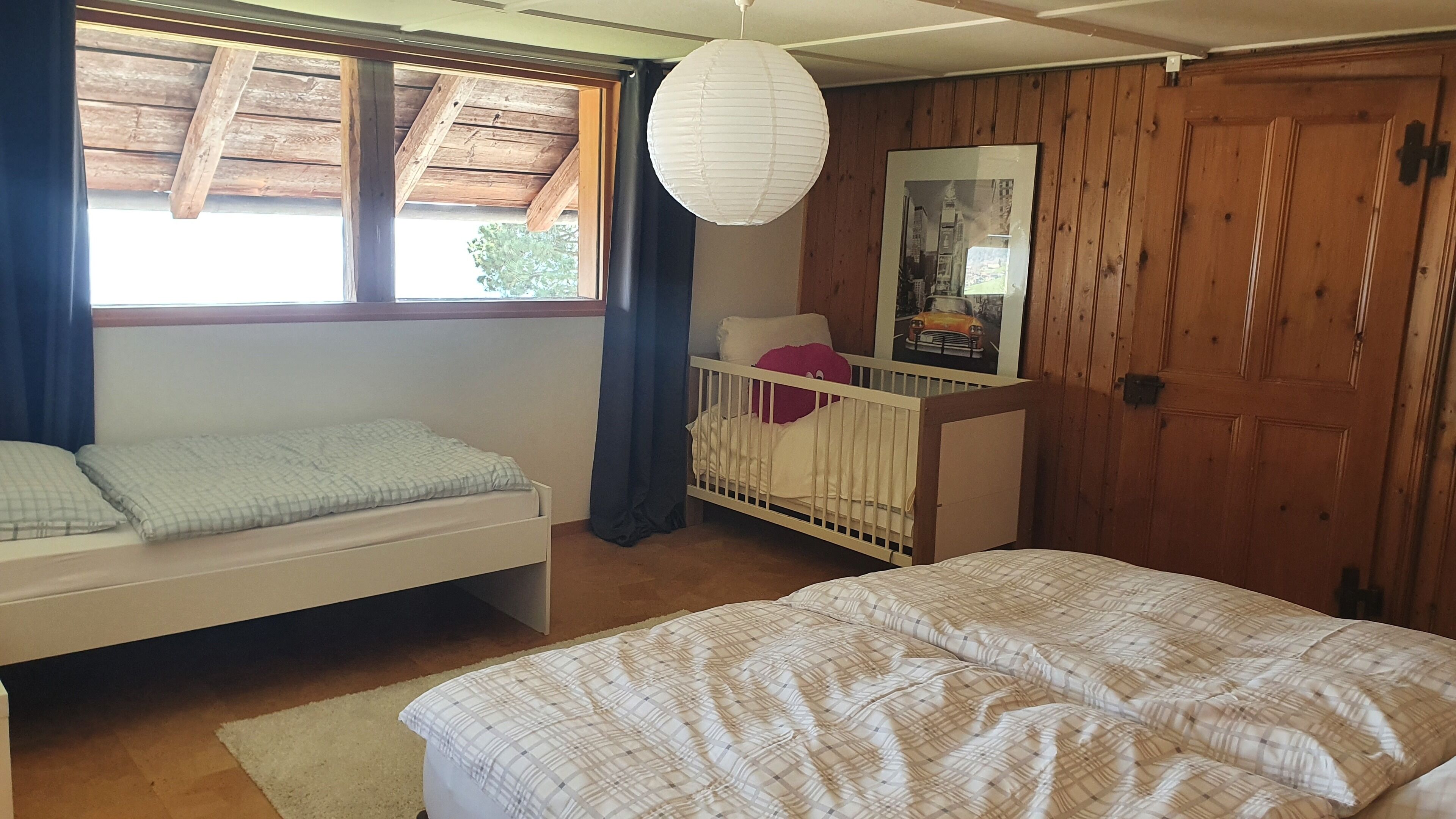 Habitació amb un llit, una bressol, una finestra amb cortines, un sostre de fusta i un quadre penjat a la paret.