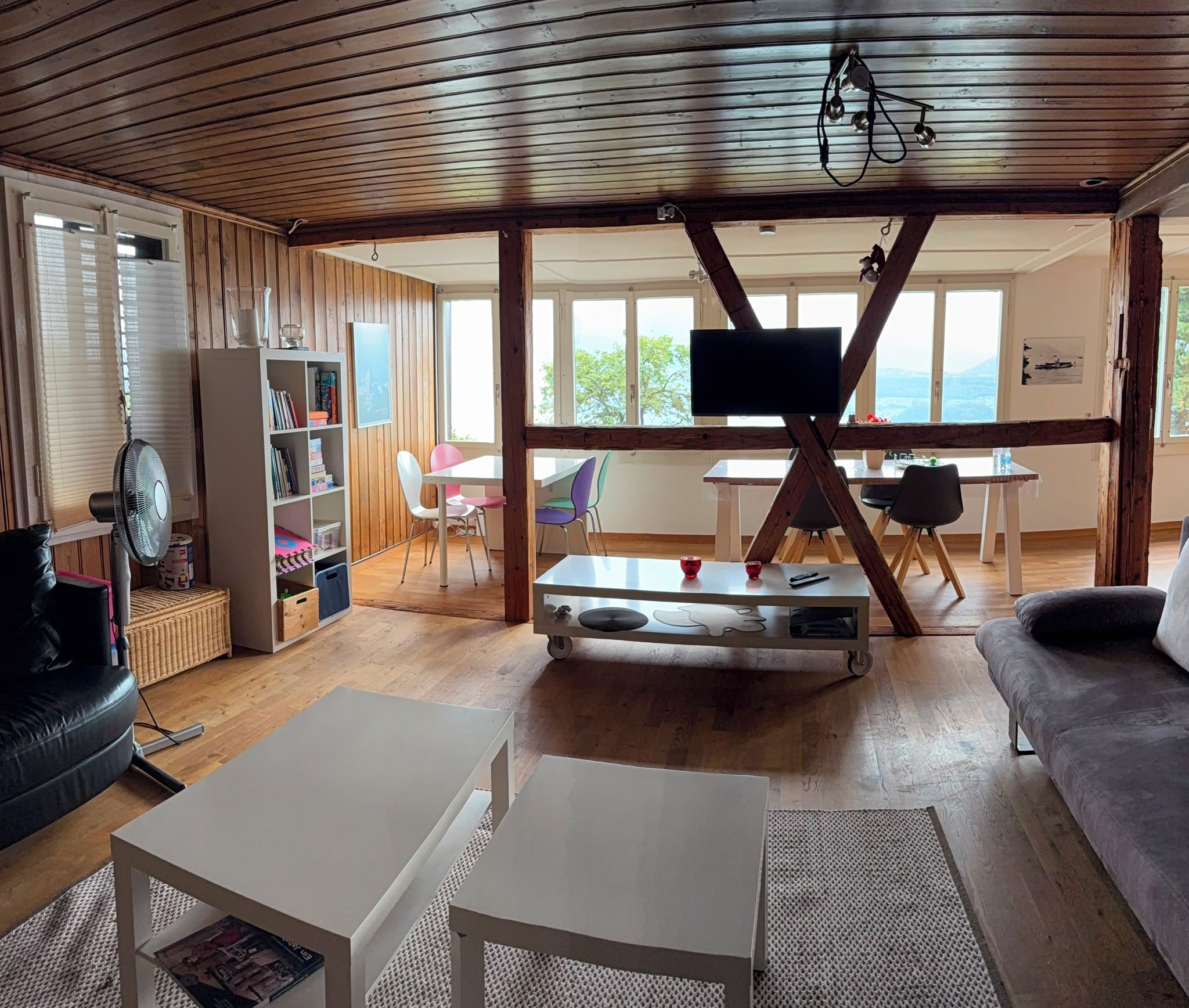 Un vaste espace de vie doté d’un plafond en bois, d’un téléviseur fixé sur une poutre en bois, d’une table blanche, d’un canapé et d’une bibliothèque.