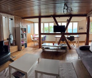 A spacious living area with a wooden ceiling, a television mounted on a wooden beam, a white table, a sofa, and a bookshelf. - Comfortable vacation home overlooking Lake Thun and the Alpine chain (Sigriswil)
