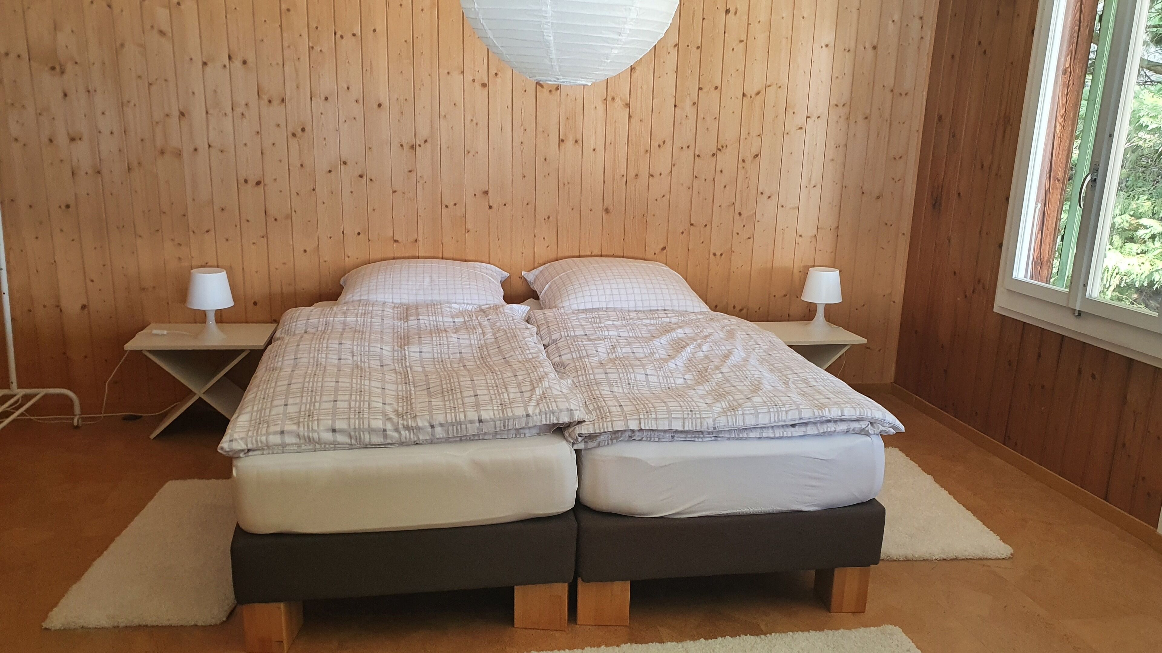 A double bed with a plaid duvet, two bedside tables with lamps, wooden walls, and a window with a view of greenery.