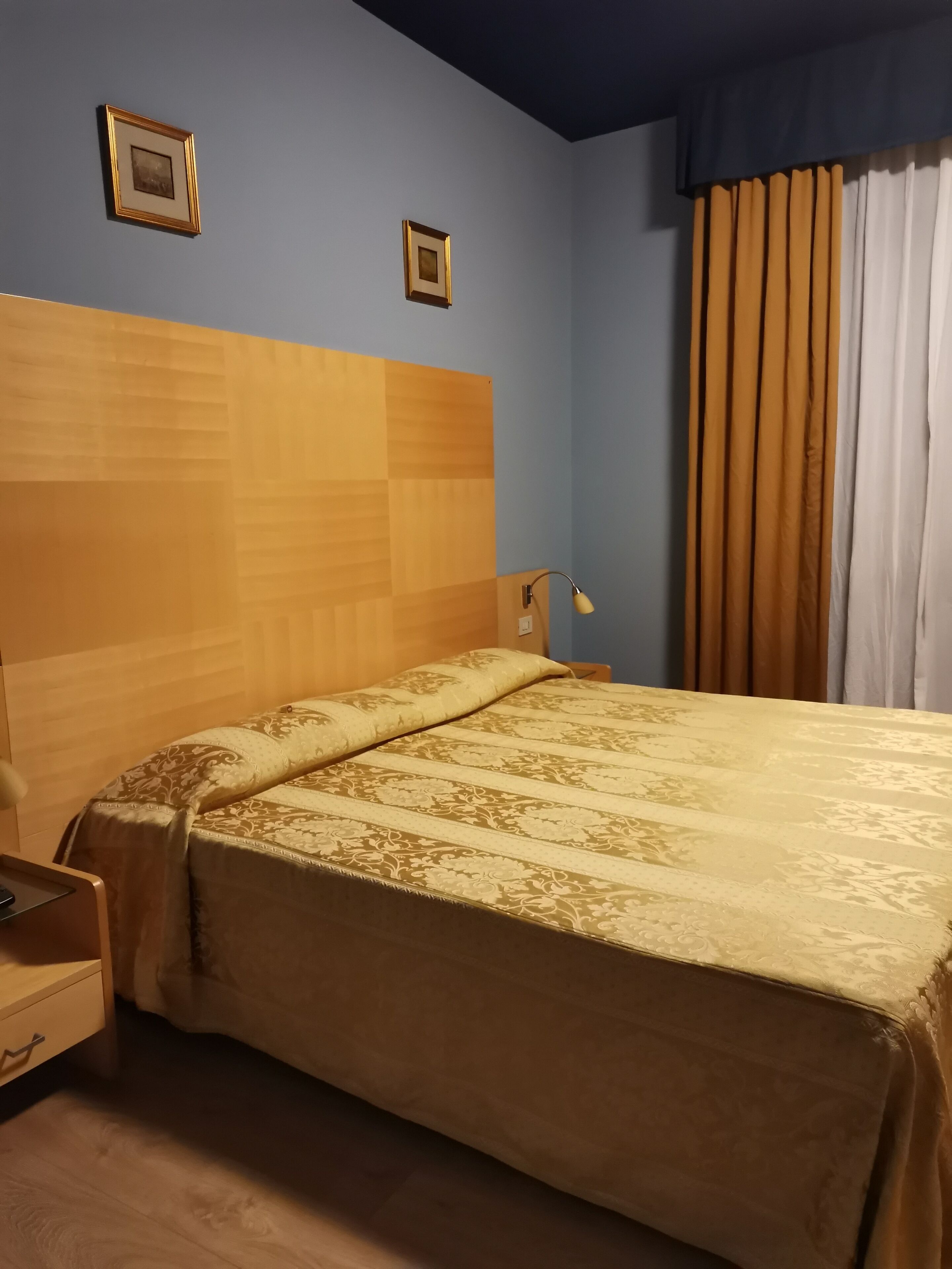 A hotel room with a wooden headboard, a bed with a patterned bedspread, a nightstand, a lamp, and a window with curtains.