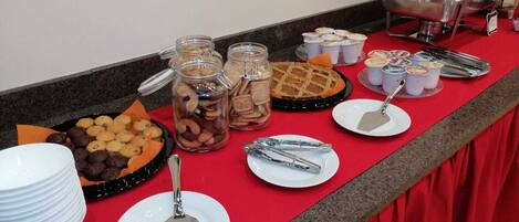 Taula de buffet amb diversos aliments, com ara galetes, pastissos i una taronja, juntament amb els utensilis per servir i recipients.