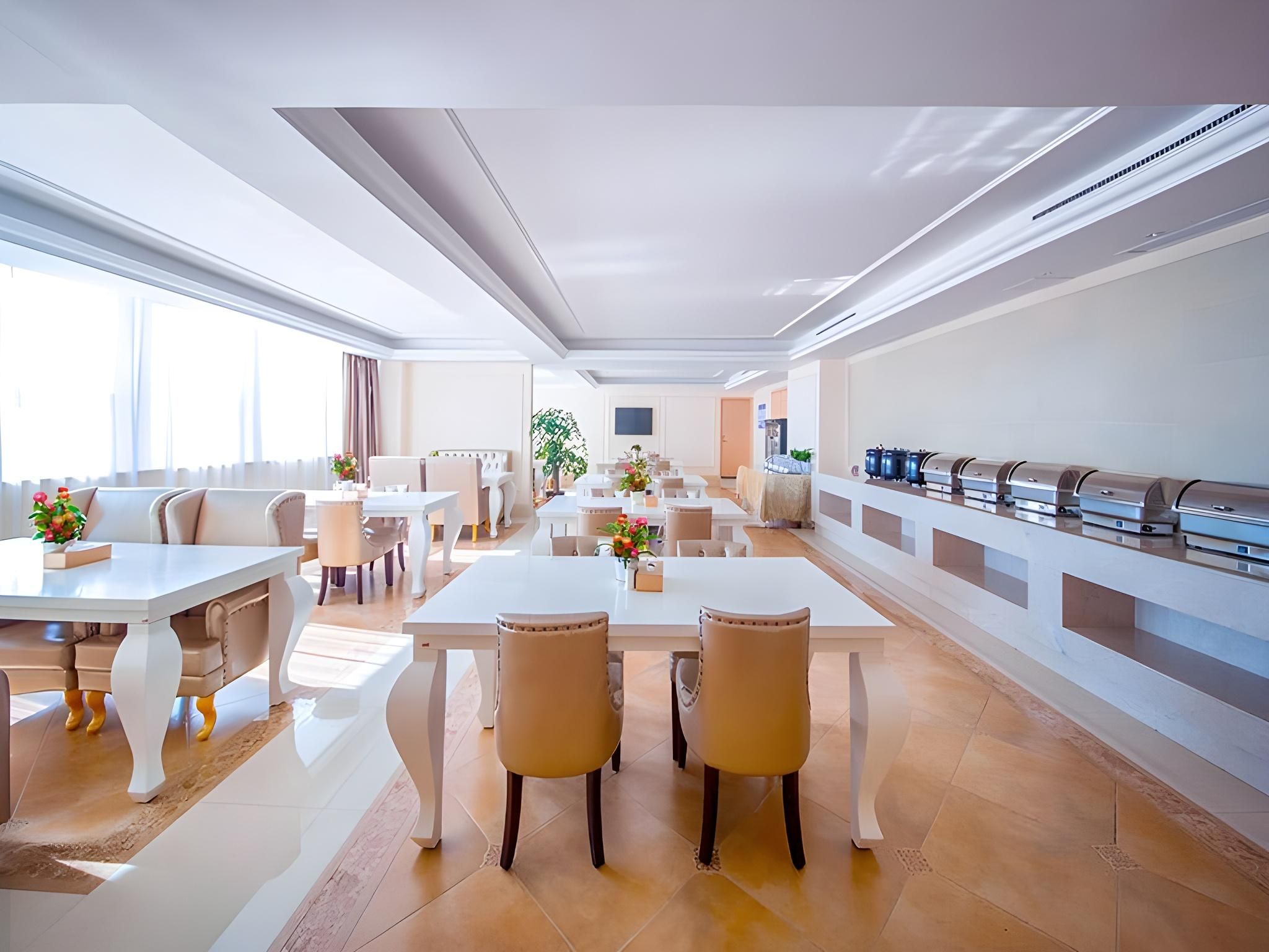 Un comedor de hotel moderno con varias mesas y sillas, una barra de buffet y un amplio ventanal que proporciona luz natural.