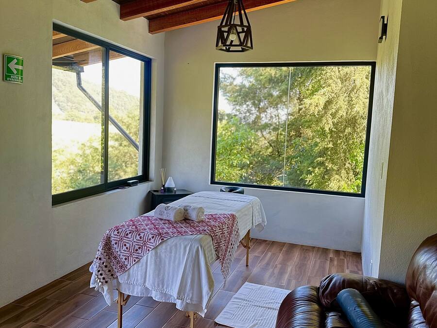 Una habitación con cama, piso de madera, sofá de cuero y ventanas amplias con vista a árboles.