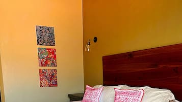 Una cama con colcha blanca y rosa, dos almohadas y un cabecero de madera. Encima de la cama, en la pared, hay tres cuadros enmarcados.