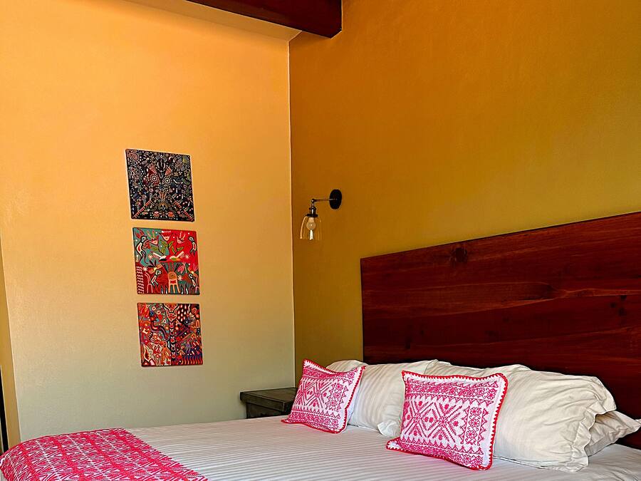 Una cama con colcha blanca y rosa, dos almohadas y un cabecero de madera. Encima de la cama, en la pared, hay tres cuadros enmarcados.